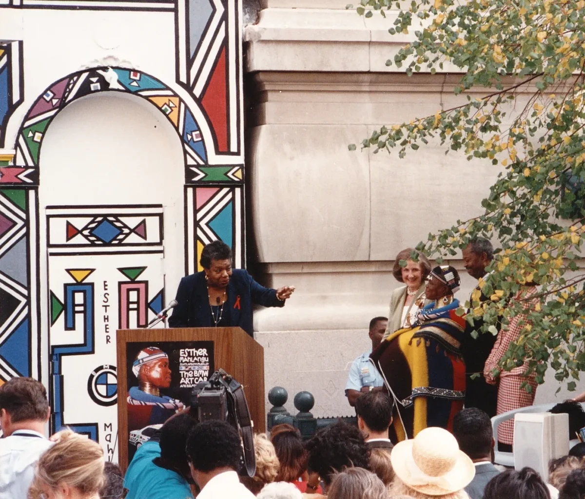 A dark-skinned woman stands at a podium which is set up outside of a large building and in front of a large crowd of people. The woman points to another dark-skinned woman to the right, who is dressed in colorful, traditional African dress and jewelry. She smiles warmly next to a light-skinned woman with shoulder-length brown hair and a dark-skinned man. Behind the podium is a large geometric mural.