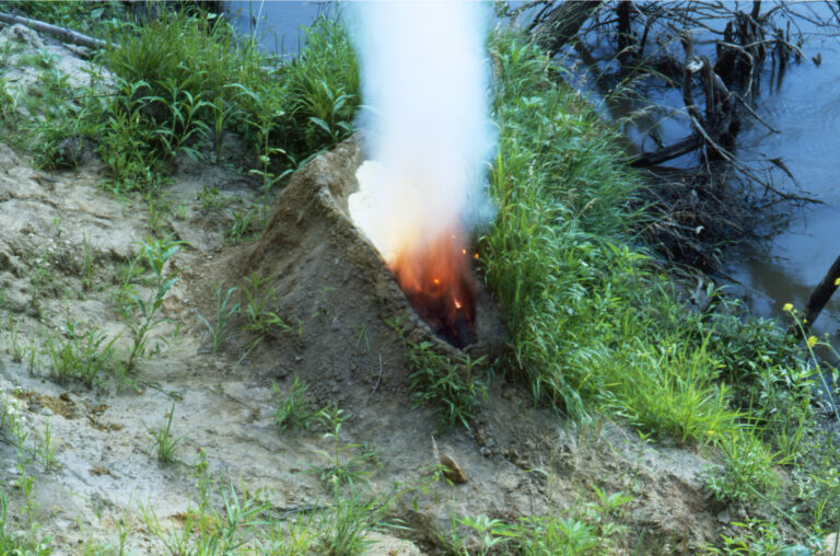 A mound of dirt in a grassy area by water. In the center of the mound is a human-shaped recess full of gunpowder that has been set aflame, exploding up in fire and white smoke, making the dirt mound resemble an erupting volcano.