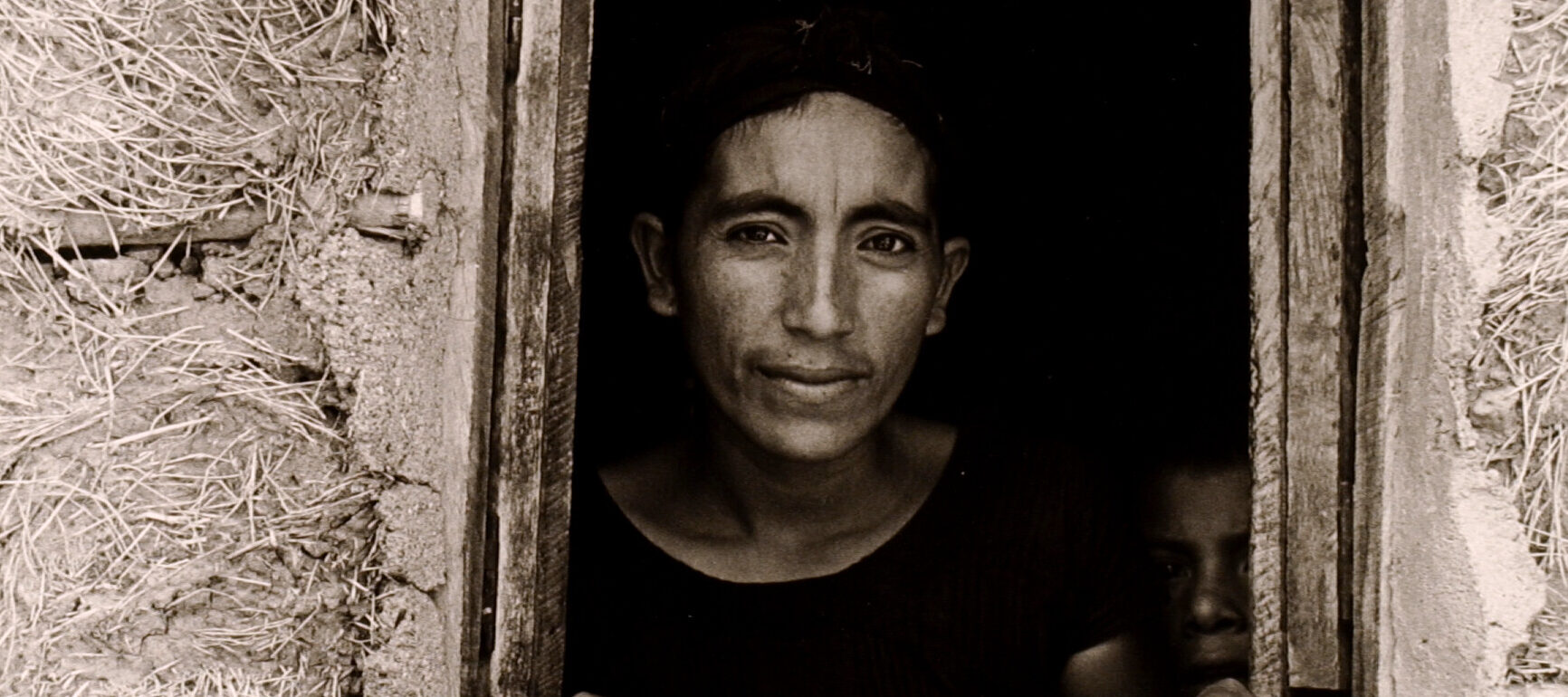 A black-and-white photograph features an open, rough-hewn window framing the head and shoulders of a woman with dark hair and medium-dark skin. Peering out from a murky interior, she displays 2 fish per hand on the window ledge. A mud-grass mixture textures surrounding walls.