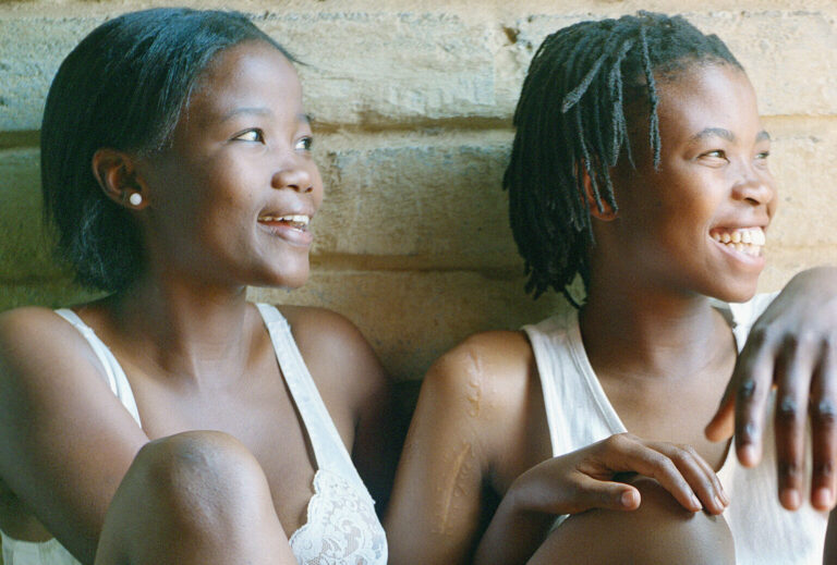 Two dark-skinned young adults with short hair sit together against a white brick wall, their limbs lovingly intertwined and glowing smiles across their faces. One wears a white bra and shorts, and the other a white tank-top and blue shorts.