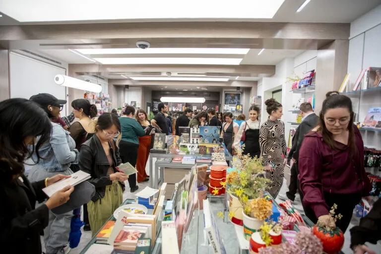 A crowded museum shop shows people browsing and buying gifts. Colorful books, plants, and trinkets fill display tables, while shelves line the walls. The atmosphere is lively and bustling.