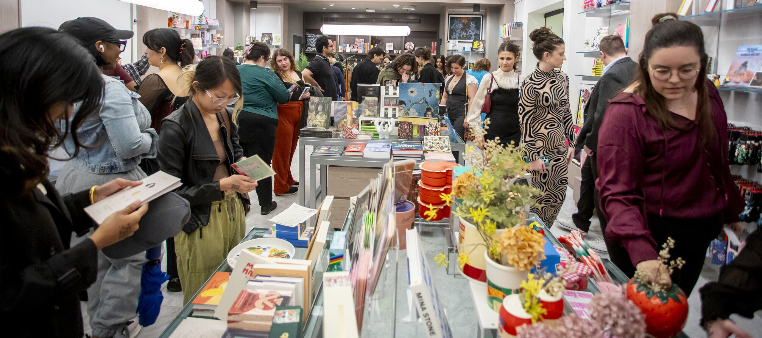 A crowded museum shop shows people browsing and buying gifts. Colorful books, plants, and trinkets fill display tables, while shelves line the walls. The atmosphere is lively and bustling.