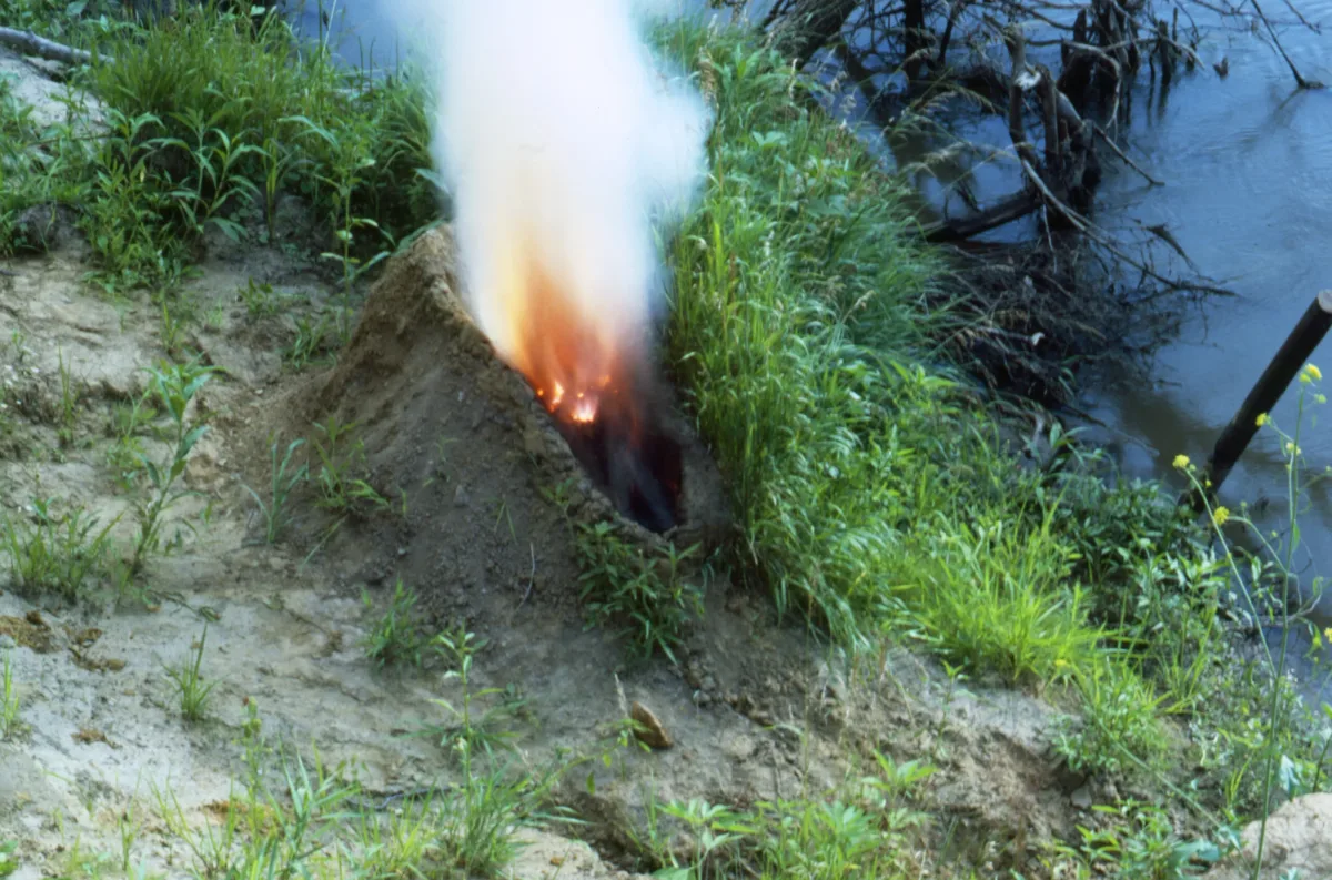 A mound of dirt in a grassy area by water. In the center of the mound is a human-shaped recess full of gunpowder that has been set aflame, exploding up in fire and white smoke, making the dirt mound resemble an erupting volcano.