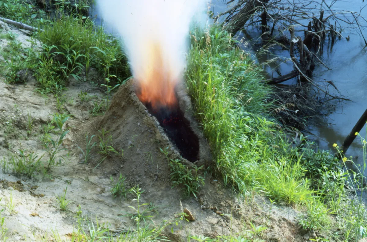 A mound of dirt in a grassy area by water. In the center of the mound is a human-shaped crater full of gunpowder that has been set aflame, exploding up in fire and smoke, making the dirt mound resemble an erupting volcano.
