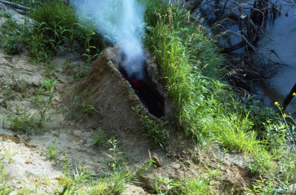 A mound of dirt in a grassy area by water. In the center of the mound is a human-shaped recess full of gunpowder that has been set aflame, exploded, and now the remaining ashes billow smoke into the air.