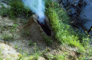 A mound of dirt in a grassy area by water. In the center of the mound is a human-shaped recess full of gunpowder that has been set aflame, exploded, and now the remaining ashes billow smoke into the air.