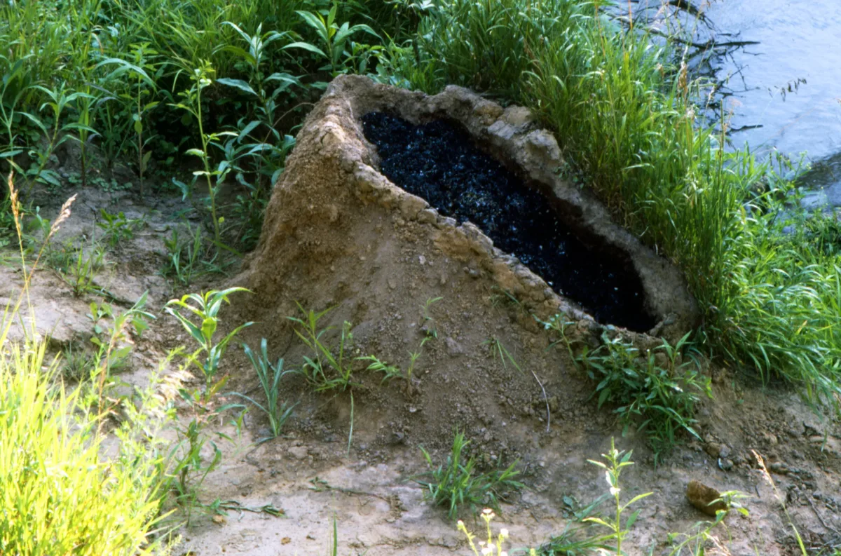 A mound of dirt in a grassy area by water. In the center of the mound is a human-shaped recess full of gunpowder that has been set aflame, exploded, and now is filled with black lifeless ashes.