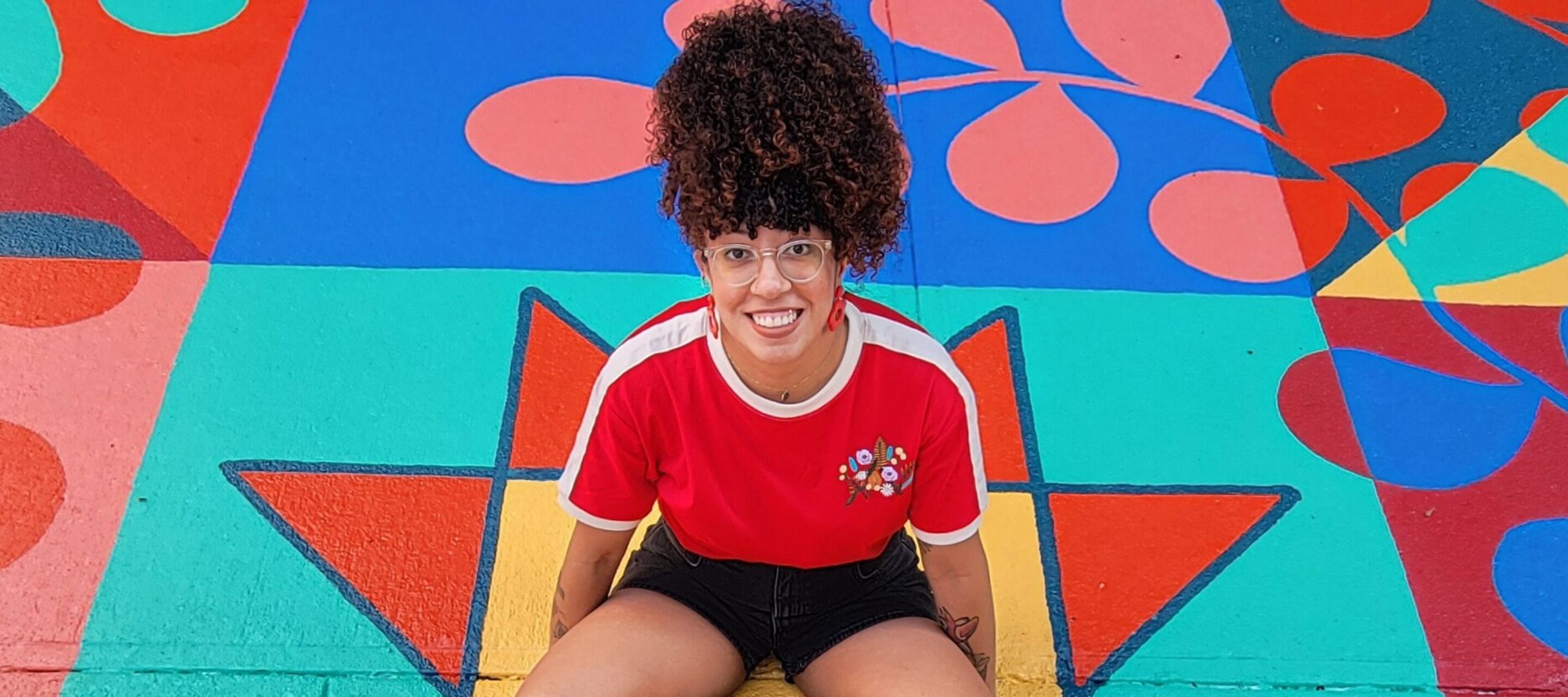 A young woman with coily, curly hair worn in a high ponytail sits in the middle of a bright mural painted on the ground. She smiles happily.