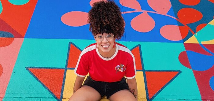 A young woman with coily, curly hair worn in a high ponytail sits in the middle of a bright mural painted on the ground. She smiles happily.