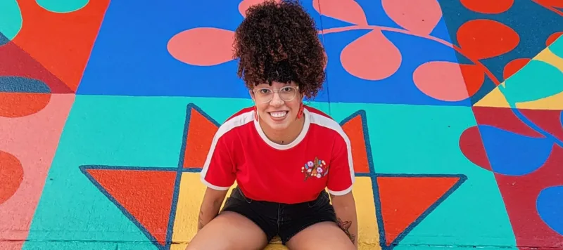 A young woman with coily, curly hair worn in a high ponytail sits in the middle of a bright mural painted on the ground. She smiles happily.