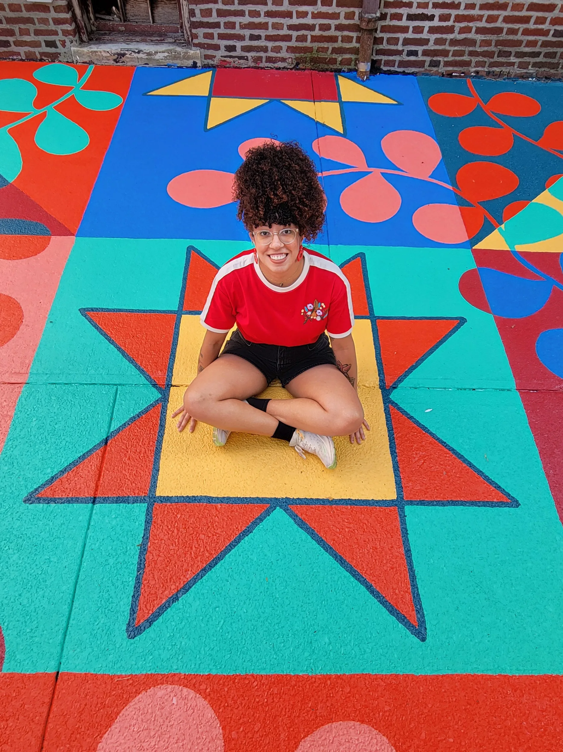 A young woman with coily, curly hair worn in a high ponytail sits in the middle of a bright mural painted on the ground. She smiles happily.
