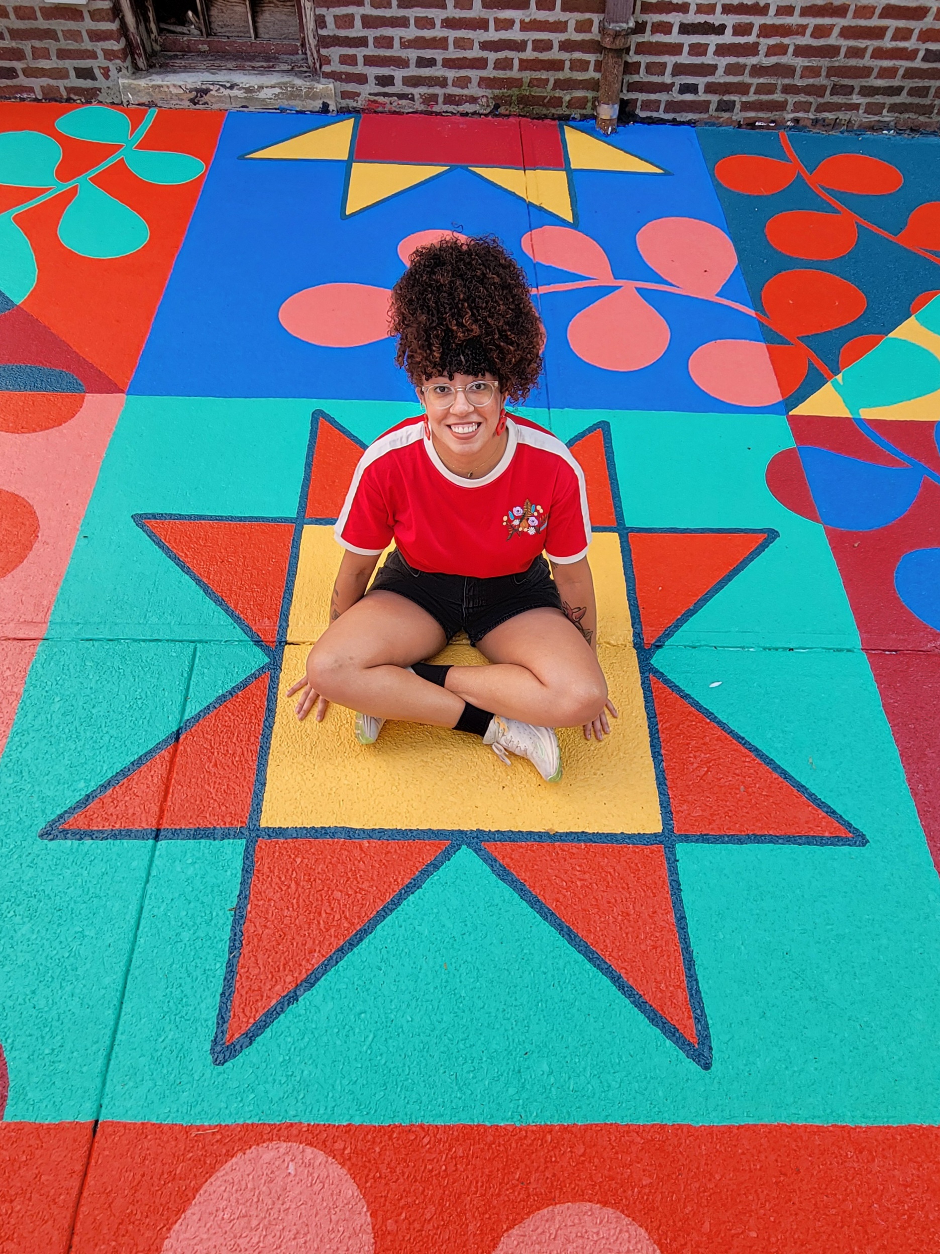 A young woman with coily, curly hair worn in a high ponytail sits in the middle of a bright mural painted on the ground. She smiles happily.