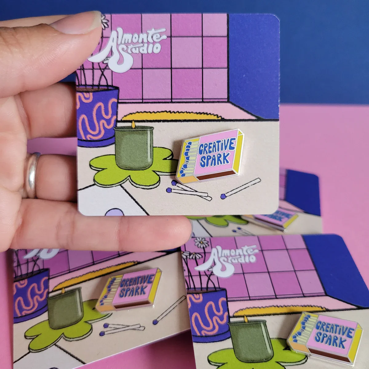 A light-skinned hand holds up a colorful enamel pin affixed to an illustrated square. The pin is in the shape of a matchbox and reads "Creative Spark."
