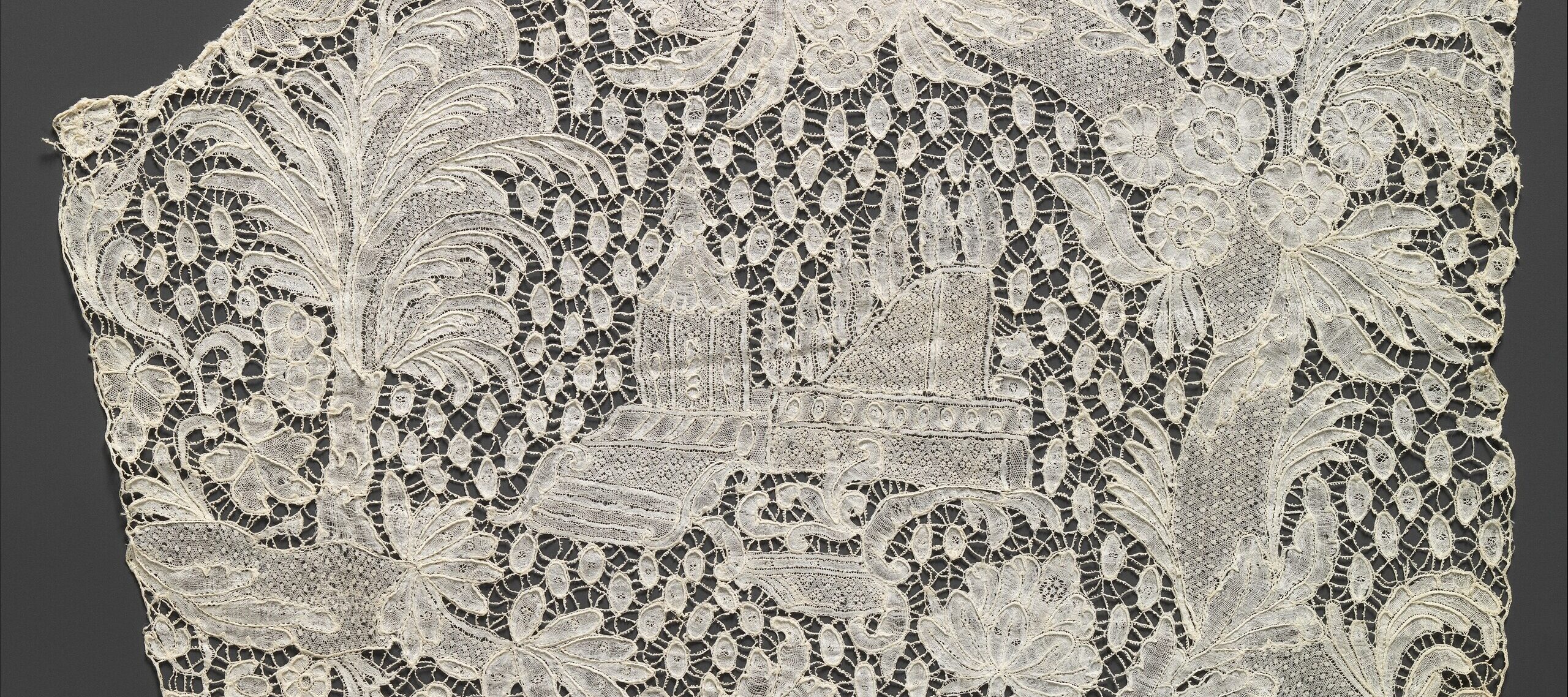 A piece of intricate, white lace features floral patterns and an embroidered scene of a building with trees in the center, set against a dark gray background.