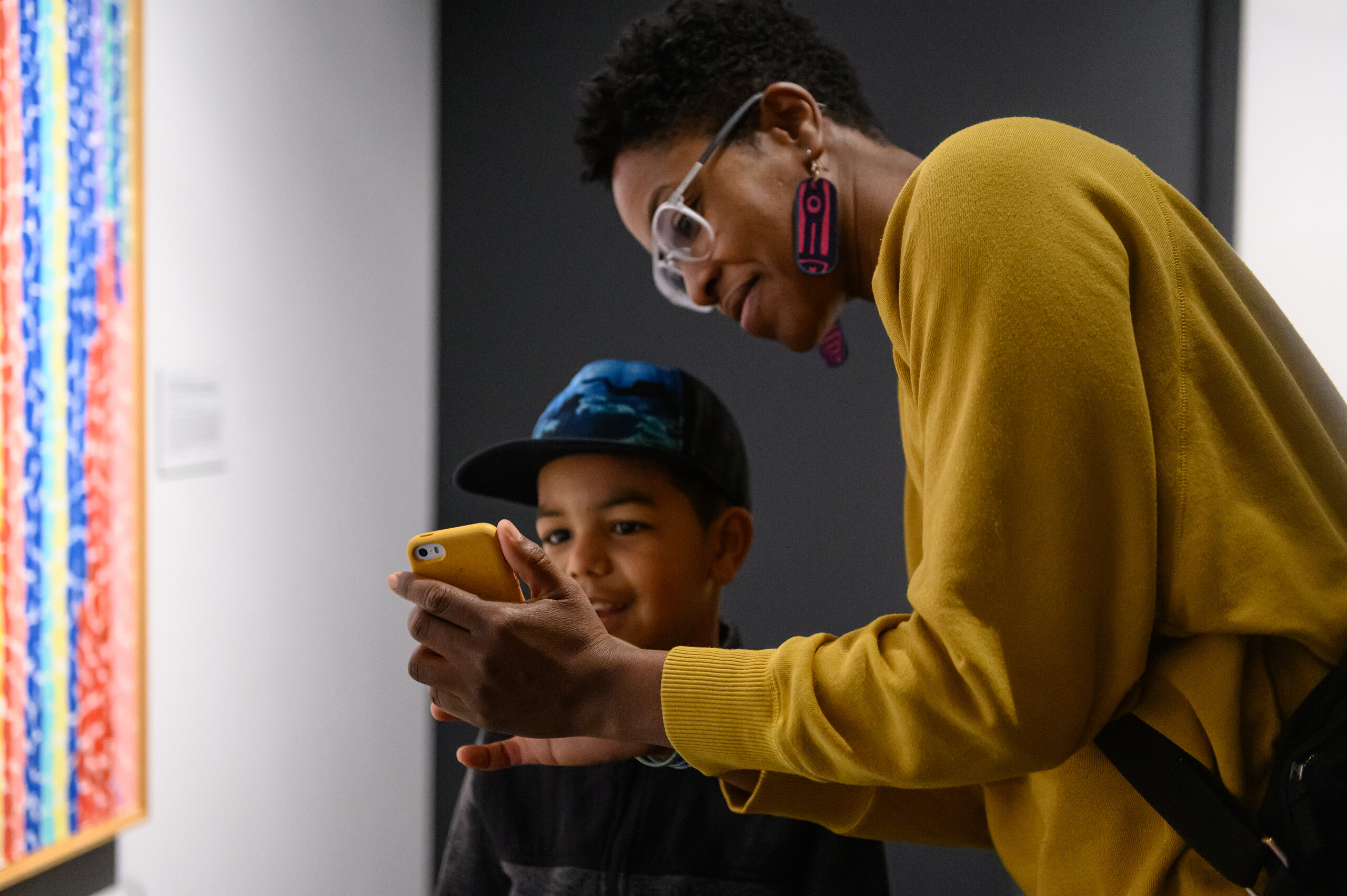A dark-skinned woman and young child stand in a museum gallery and look at a phone in front of a colorful painting.