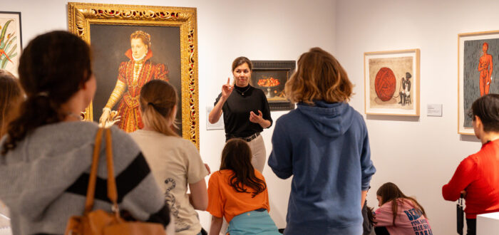 A woman with light colored skin, wearing a black top and white pants, talks to a group of people in a gallery room. She stands in front of a large, painted portrait of a woman in a high-collared red dress.