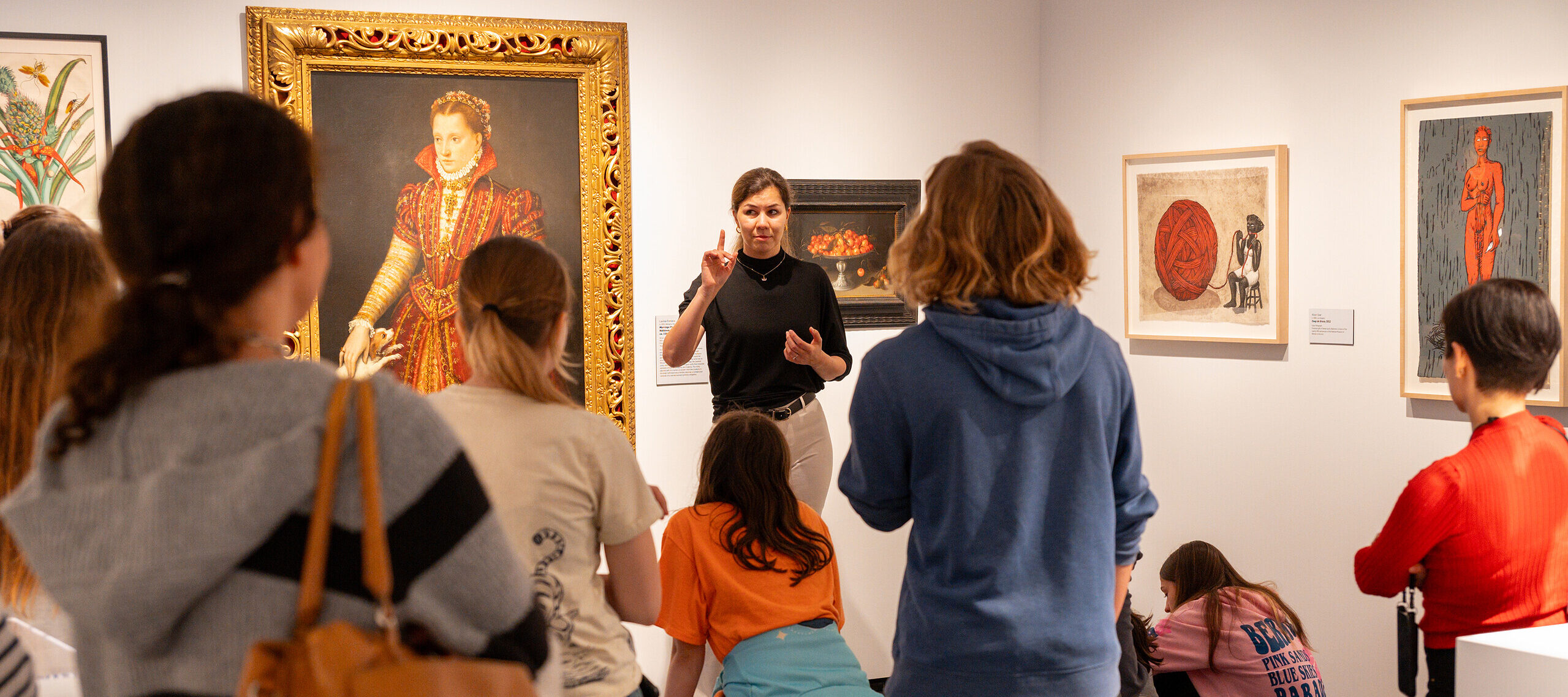A woman with light colored skin, wearing a black top and white pants, talks to a group of people in a gallery room. She stands in front of a large, painted portrait of a woman in a high-collared red dress.