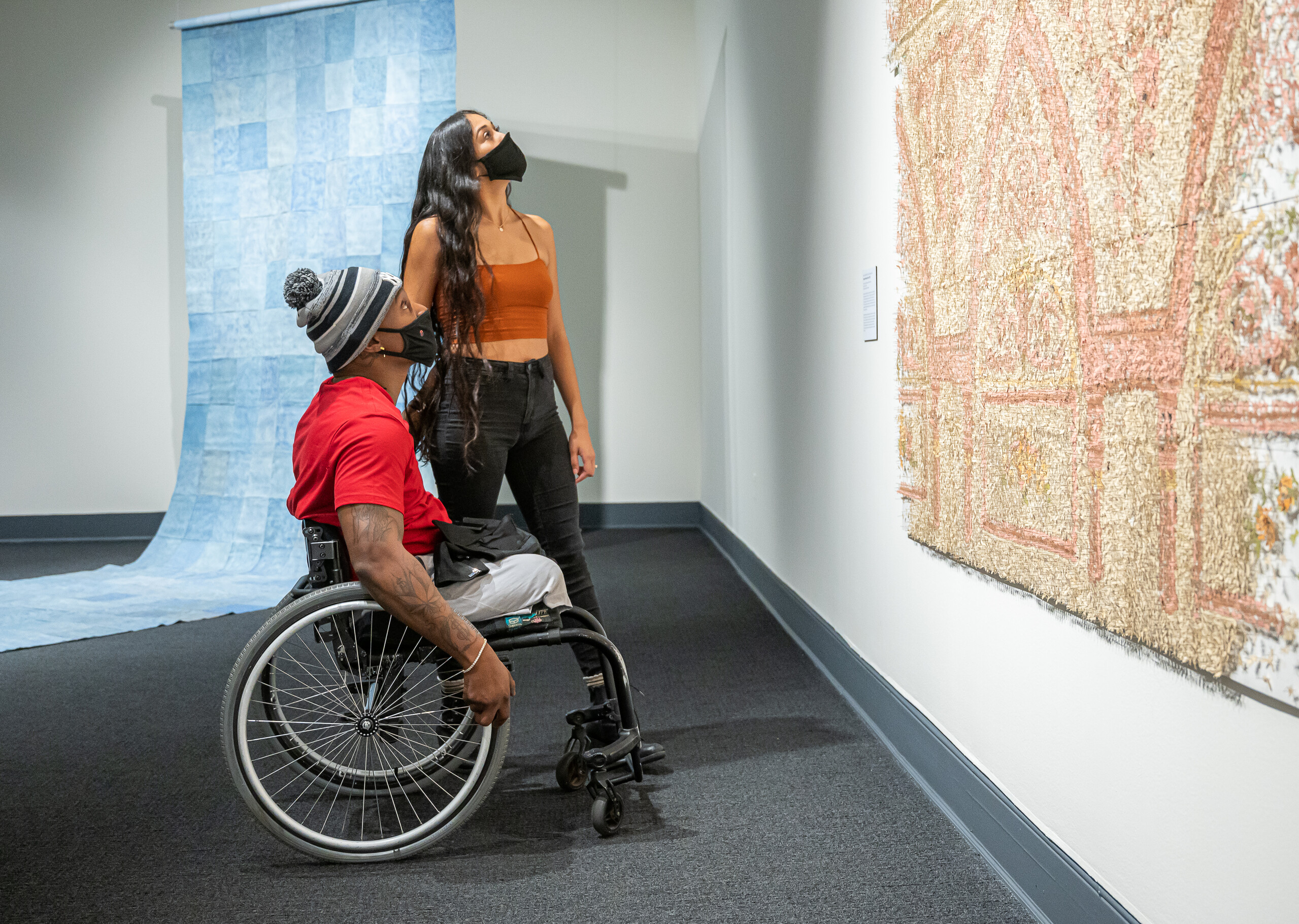 Two people look up at a large peach, tan, and ivory art installation in a gallery. The person with medium skin tone and tattoos on their right arm sits in a wheelchair and wears a red t-shirt, gray pants, gray-and-black striped winter hat, and black face mask. A person with medium skin tone, long curly black hair stands behind, wearing a burnt orange tank top, black jeans, and a black face mask. A large checkered blue artwork unfolds onto the gallery floor behind the pair.