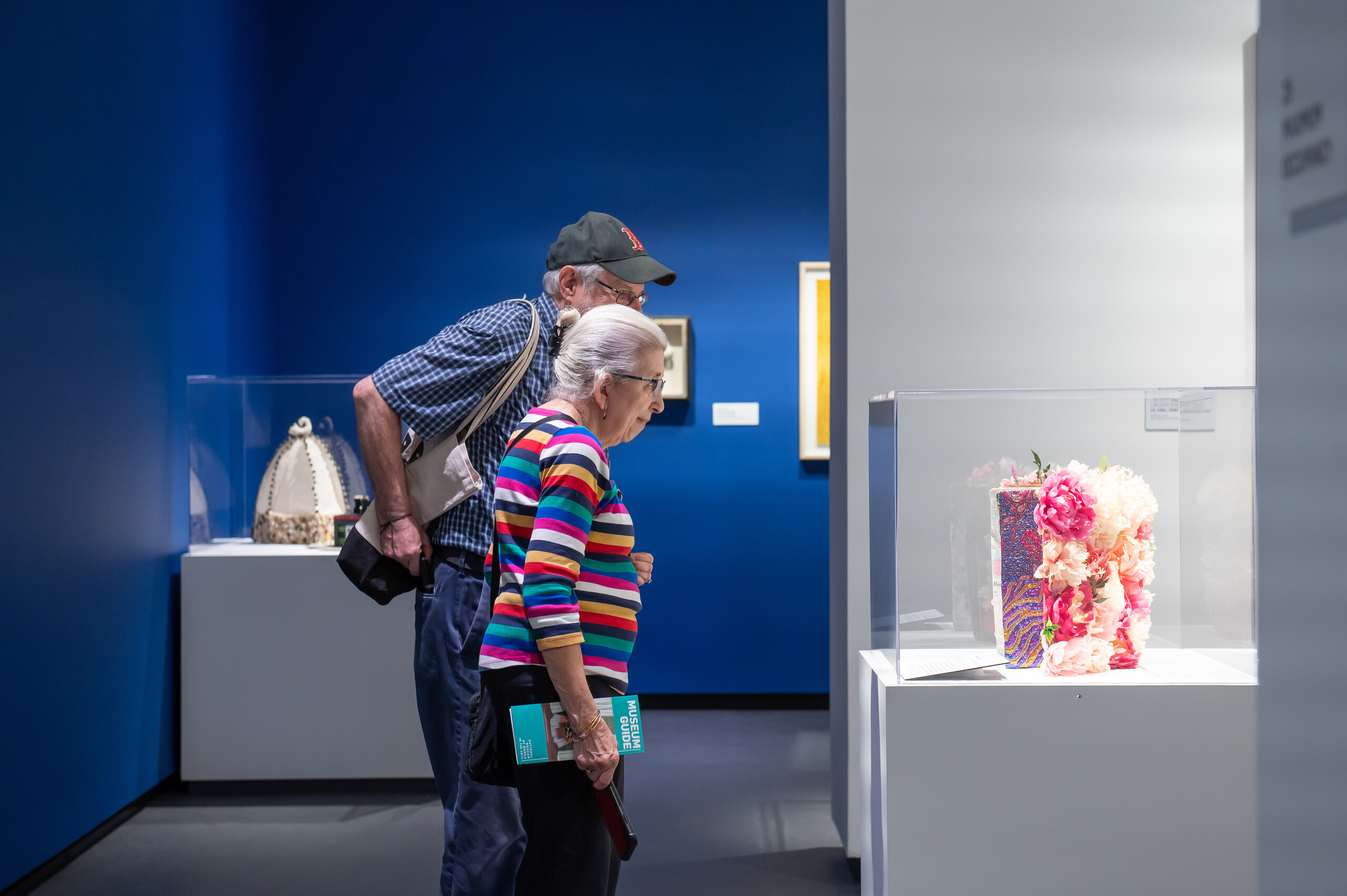 An older light-skinned man and woman look at a sculptural book on display in a glass case in a museum gallery with royal blue walls. Pink and white flowers bloom from the back cover of the book and glittery designs appear on one of the pages.