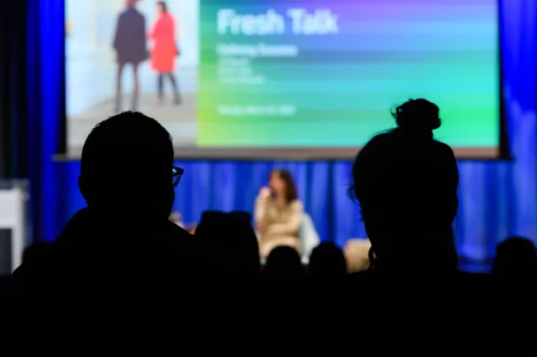 Audience in silhouette watches a seated speaker on stage. A colorful presentation screen in the background displays text including "Fresh Talk."