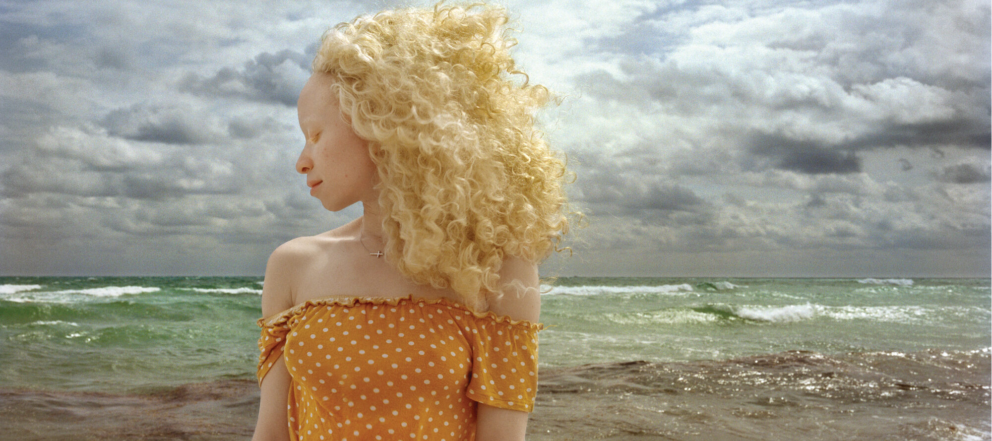 In front of a stormy ocean, a woman with light skin and blonde, wind-blown hair stands with head turned in profile and eyes closed.