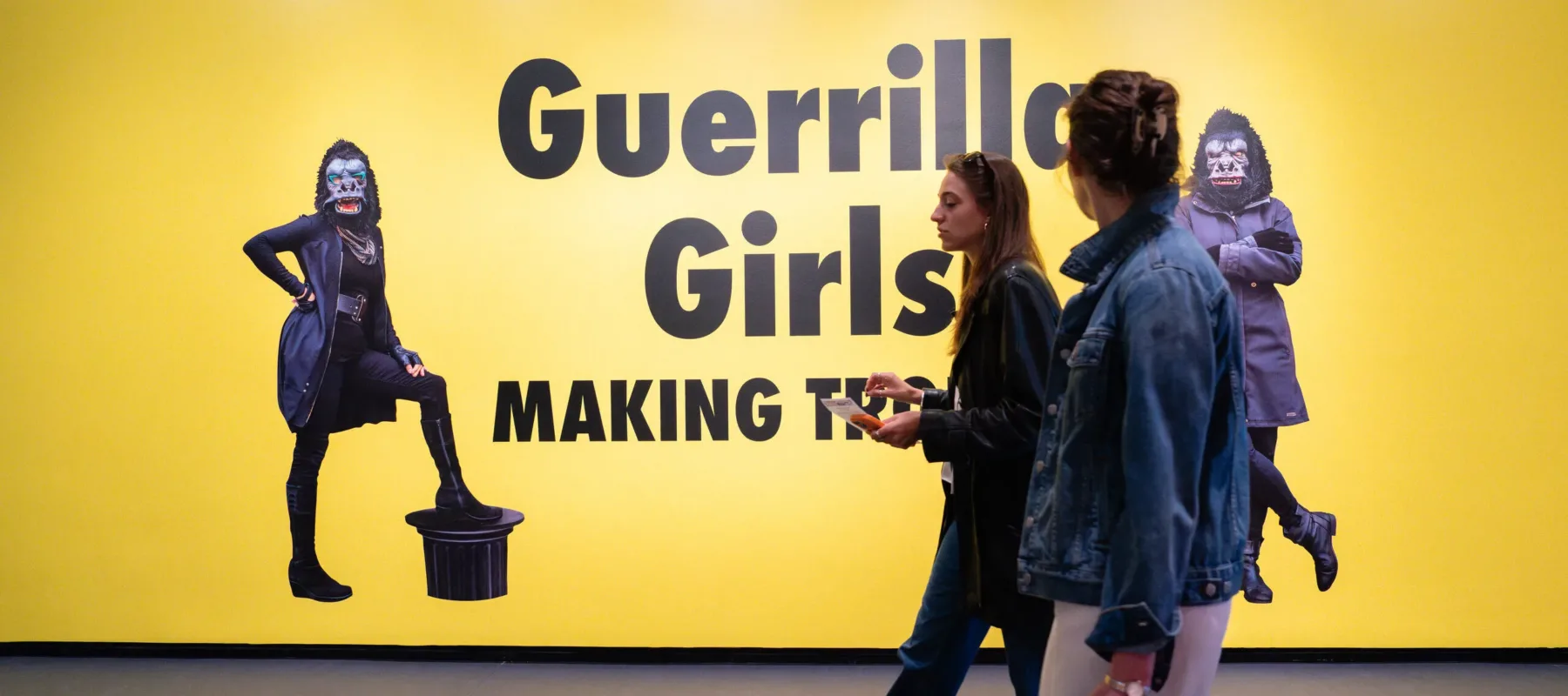 Two women walk past a bright yellow wall with large text that reads "Guerrilla Girls MAKING TRB" and images of masked figures in black clothing and gorilla masks standing on either side.