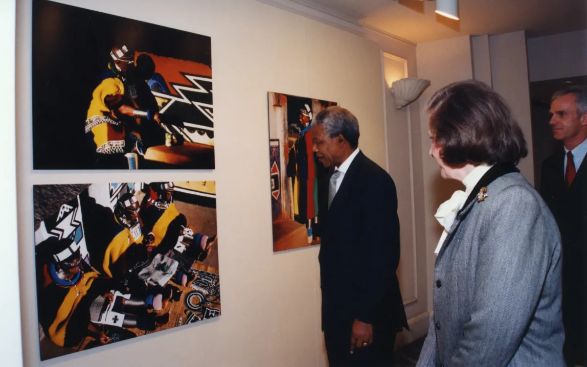 Three people in formal attire view colorful, abstract artwork displayed on a gallery wall. Two art pieces show people in vibrant, patterned clothing. The group stands attentively, observing the art.