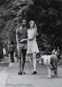 A black and white photo of a woman with light skin and a man with dark skin with their arms around each other while walking their dog in a park.