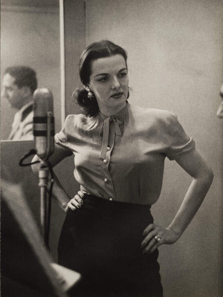 A black and white photo of a woman with light skin and dark hair. She wear s a light colored blouse and a black skirt and stands in front of a microphone. A man can be seen in the mirror reflection behind the woman