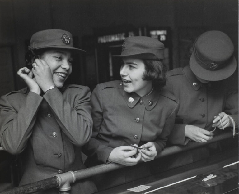 A black and white photo of three women in dark military-style uniforms wearing matching caps. The woman on the left adjusts an earring while the woman in the center speaks to her. A third woman's face is concealed by her cap as she examines her bracelet.