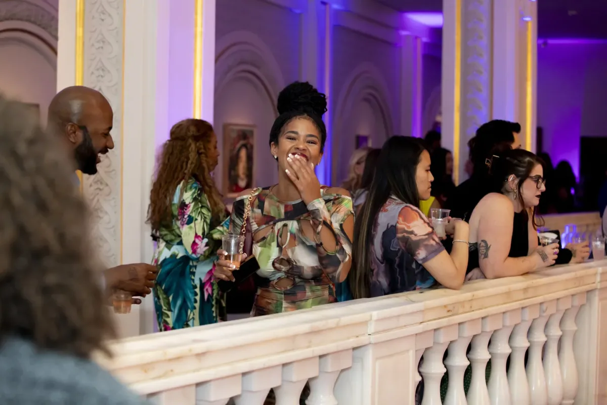A group of people stand along an ornate white balcony at a lively indoor event. One woman in the center smiles and laughs, holding a drink, while others around her chat and enjoy the atmosphere.