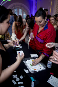 A group of women gather around a black table covered with small paper pieces, some holding drinks and interacting with the items. Ornate architecture and colorful lighting are visible in the background.