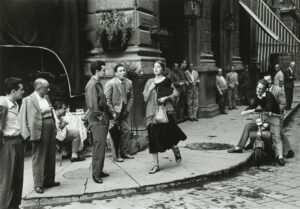 A black-and-white photograph of a young woman with light skin tone and brown hair wearing a black dress and a scarf and clutching a book and a bag walks down a city sidewalk. Several men watch her walk by, some standing against the wall of a nearby building, some sitting on chairs or motorcycles. A few men seem to be catcalling her.