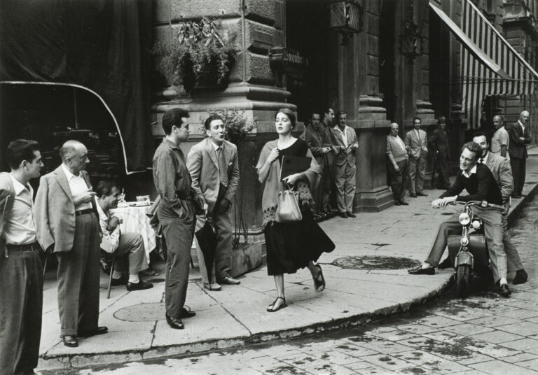 A black-and-white photograph of a young woman with light skin tone and brown hair wearing a black dress and a scarf and clutching a book and a bag walks down a city sidewalk. Several men watch her walk by, some standing against the wall of a nearby building, some sitting on chairs or motorcycles. A few men seem to be catcalling her.