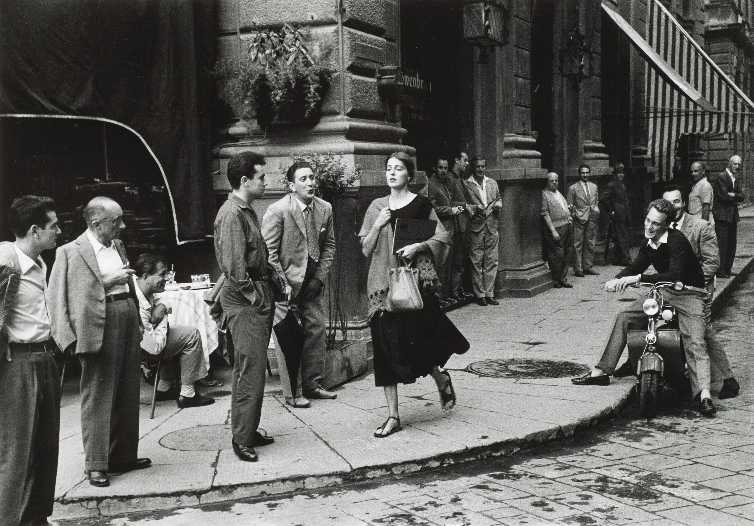 A black-and-white photograph of a young woman with light skin tone and brown hair wearing a black dress and a scarf and clutching a book and a bag walks down a city sidewalk. Several men watch her walk by, some standing against the wall of a nearby building, some sitting on chairs or motorcycles. A few men seem to be catcalling her.