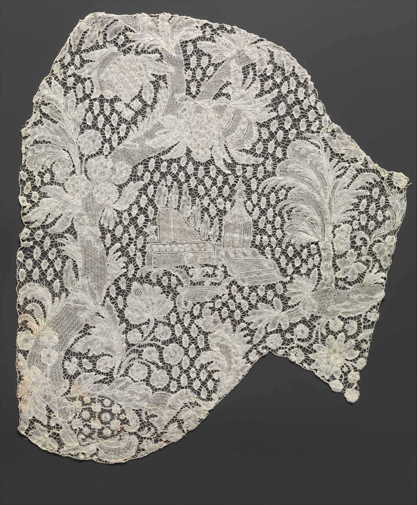 A piece of intricate, white lace features floral patterns and an embroidered scene of a building with trees in the center, set against a dark gray background.