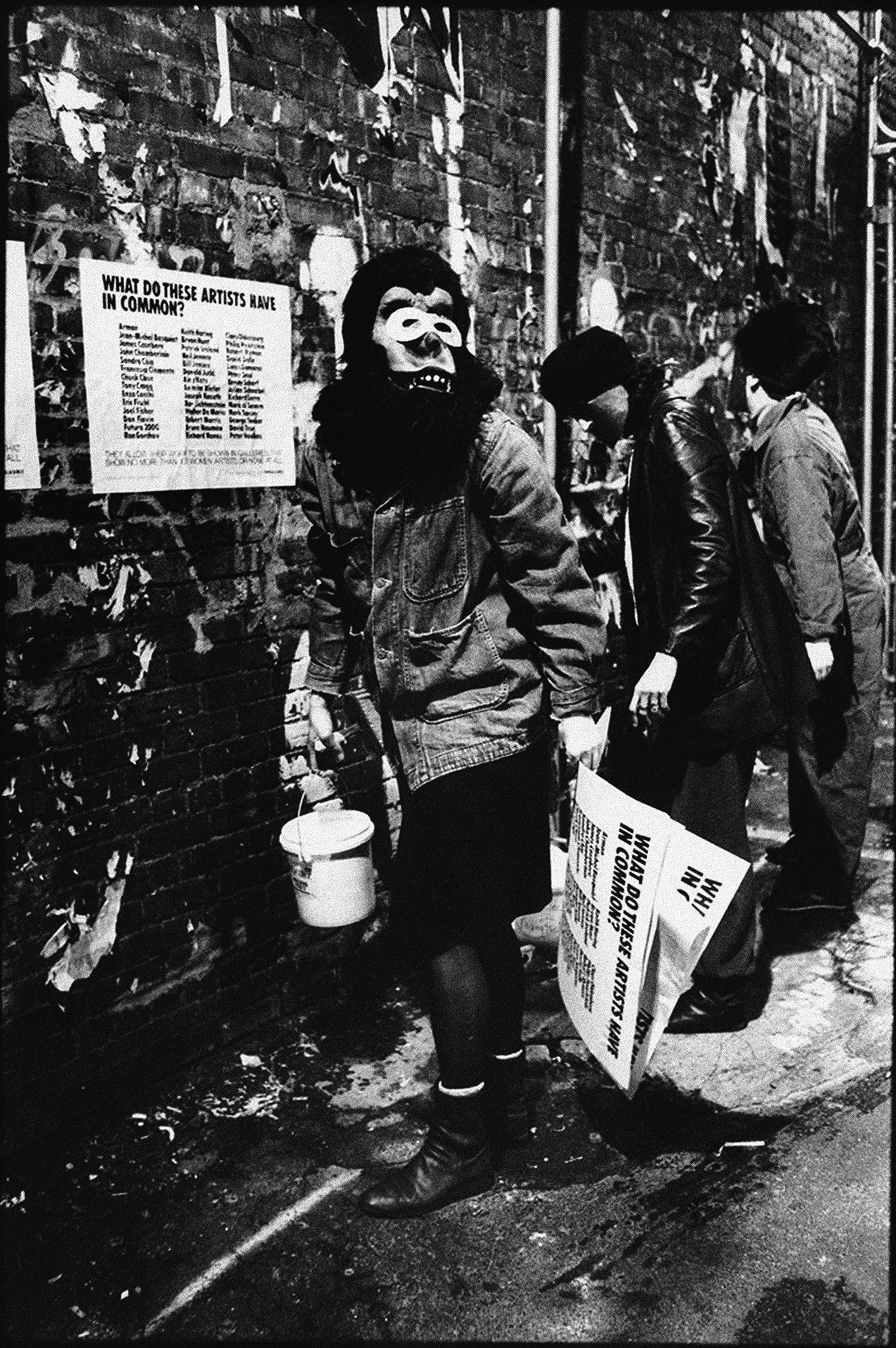A black-and-white photo shows three people standing in front of a brick wall. The person closest to the camera wears a gorilla mask, a skirt, and boots. They are holding several posters in one hand and a bucket of wheat paste in the other. One poster is already affixed to the brick wall.