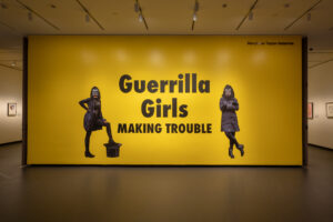 A large yellow wall with black text that says "Guerrilla Girls Making Trouble." On both sides of the text is an image of a figure wearing all black and a gorilla mask.