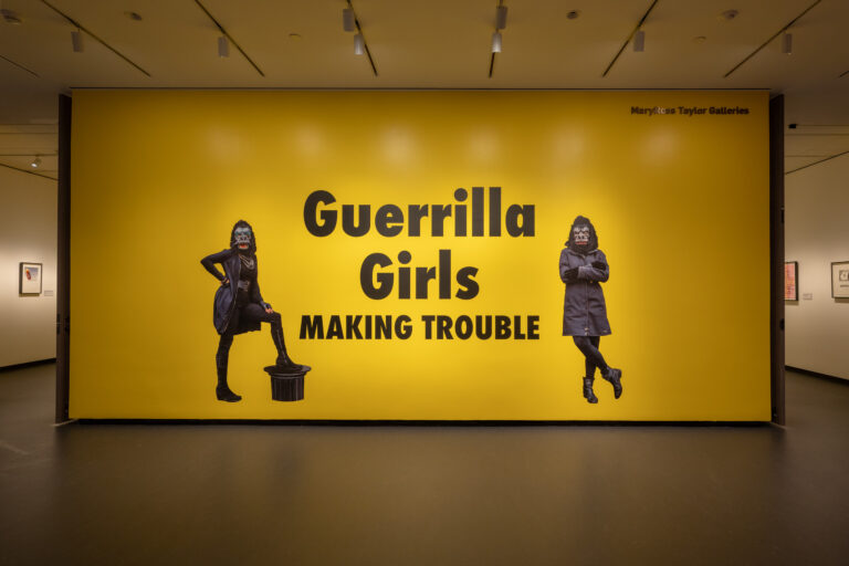 A large yellow wall with black text that says "Guerrilla Girls Making Trouble." On both sides of the text is an image of a figure wearing all black and a gorilla mask.