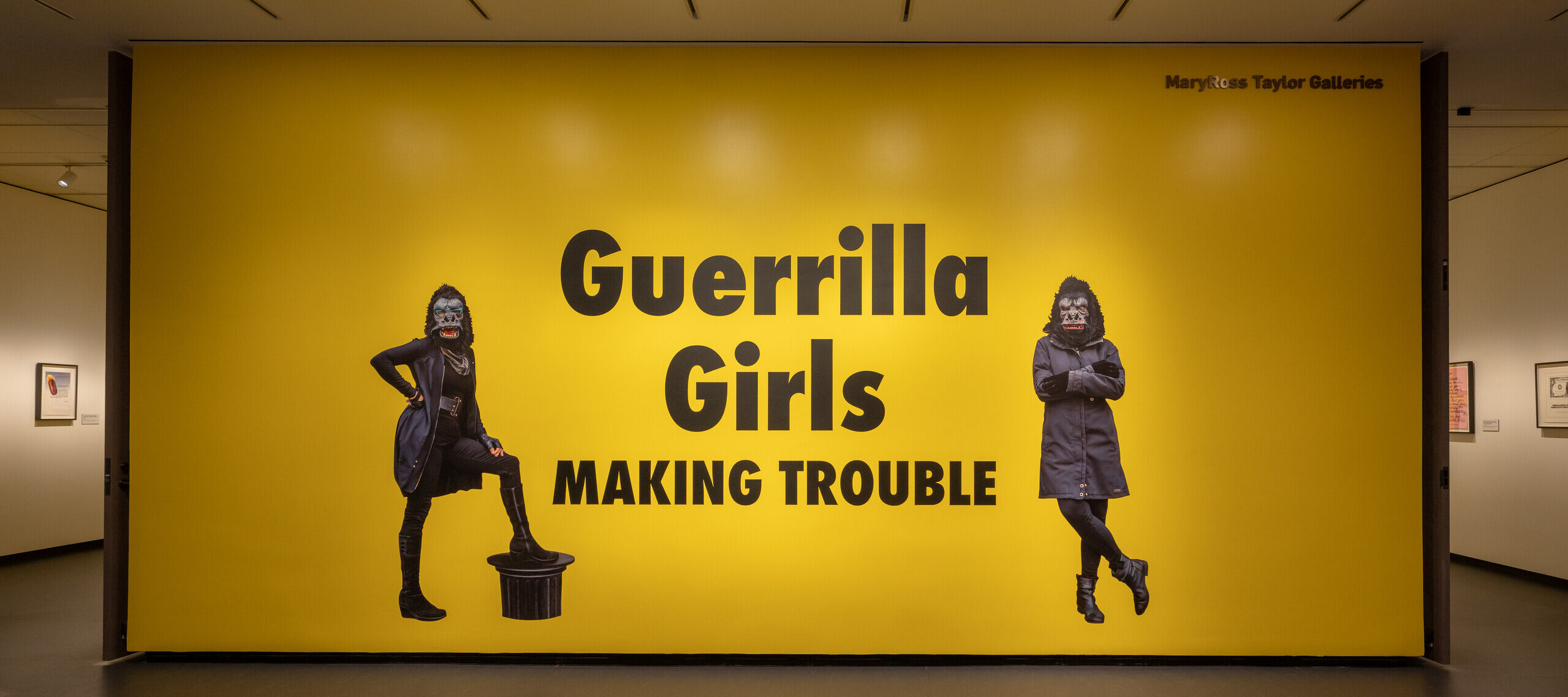 A large yellow wall with black text that says "Guerrilla Girls Making Trouble." On both sides of the text is an image of a figure wearing all black and a gorilla mask.