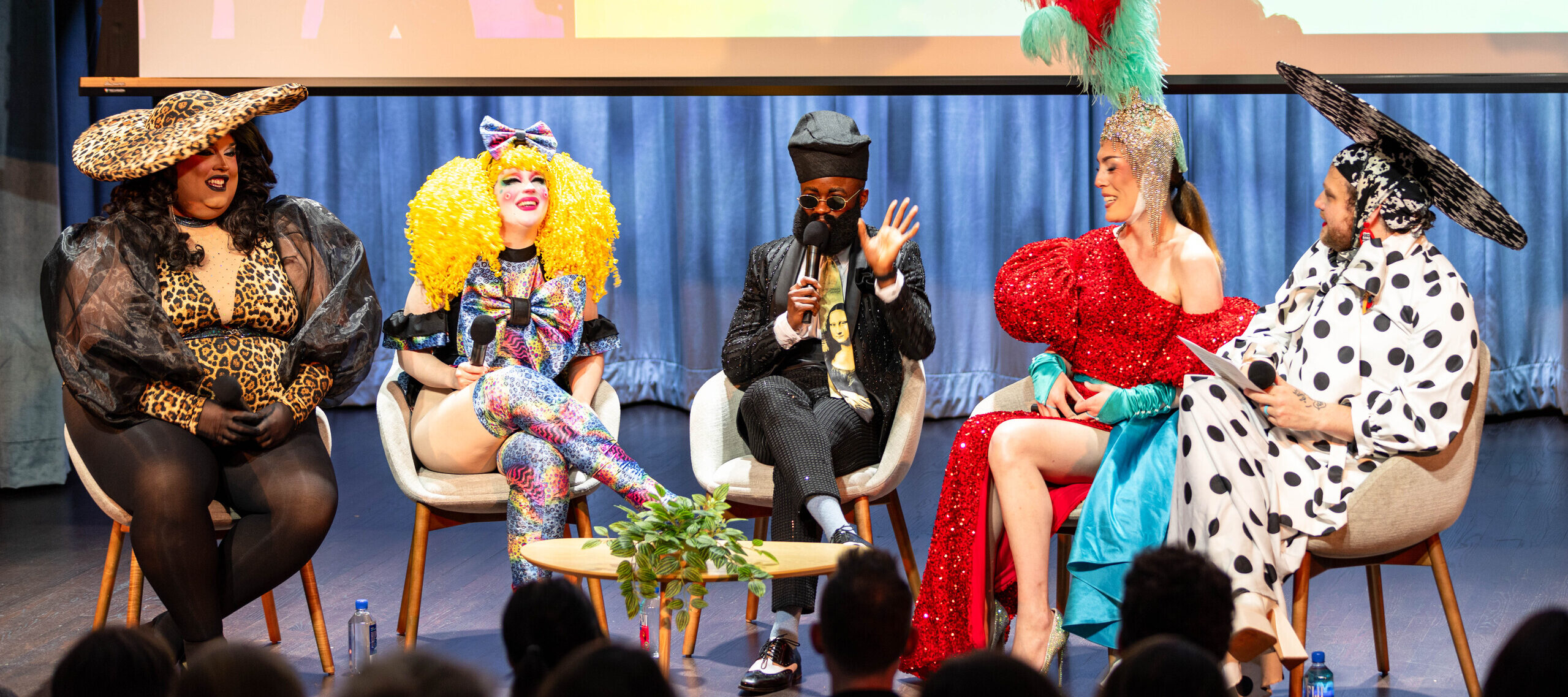 Five people in colorful, extravagant outfits and wigs sit on stage in chairs, engaged in conversation, with an audience watching. A projection screen is visible behind them.