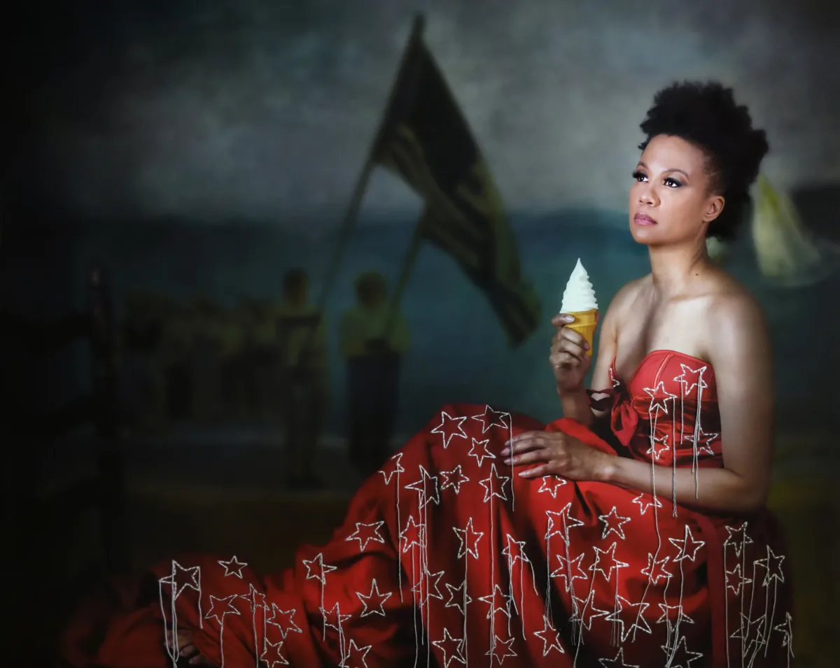 A portrait photograph of a Black woman shows her wearing a deep red formal dress with large, embroidered stars and holding an ice cream cone with a perfect conical swirl. She is seated upright, with her back to the right side of the image, looking toward the left. People holding large flags on flagpoles are visible in the dimly lit background.