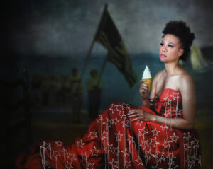 A portrait photograph of a Black woman shows her wearing a deep red formal dress with large, embroidered stars and holding an ice cream cone with a perfect conical swirl. She is seated upright, with her back to the right side of the image, looking toward the left. People holding large flags on flagpoles are visible in the dimly lit background.