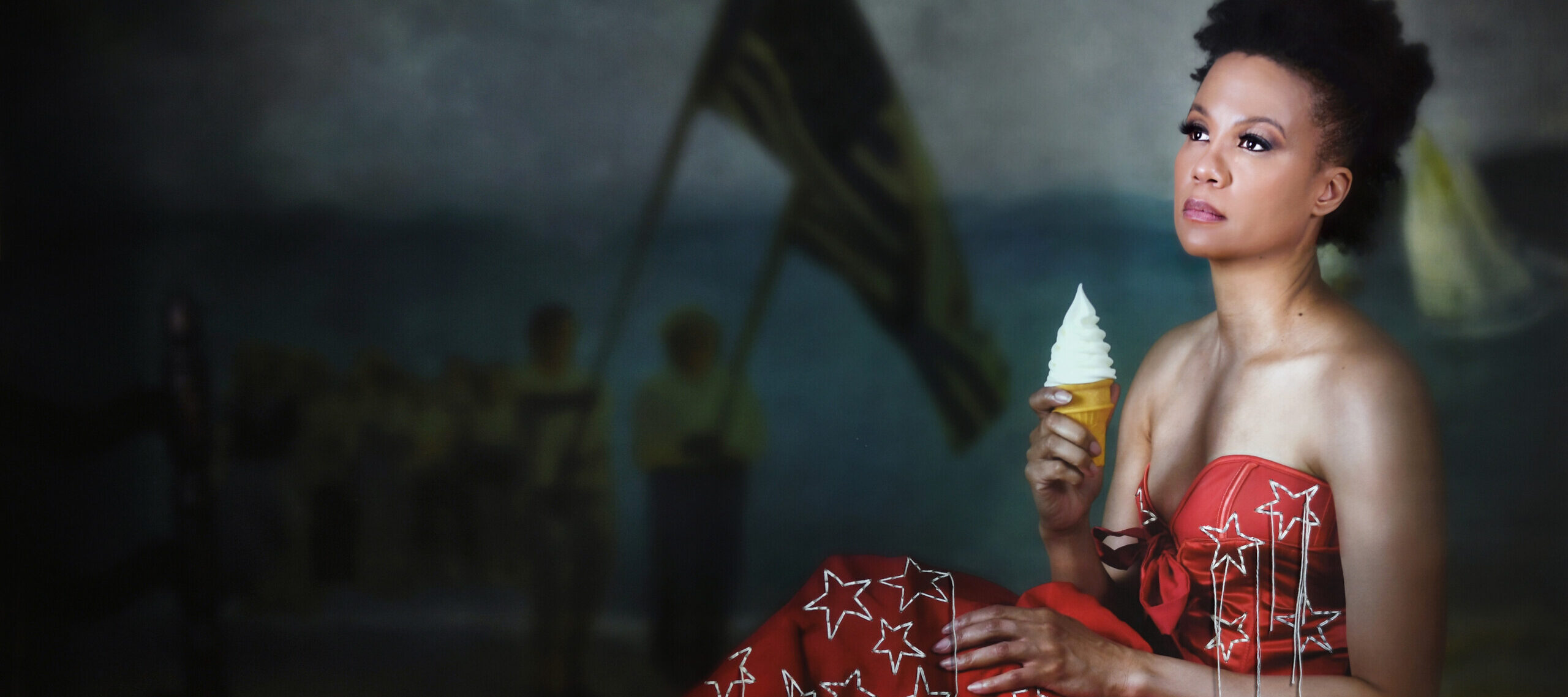 A portrait photograph of a Black woman shows her wearing a deep red formal dress with large, embroidered stars and holding an ice cream cone with a perfect conical swirl. She is seated upright, with her back to the right side of the image, looking toward the left. People holding large flags on flagpoles are visible in the dimly lit background.