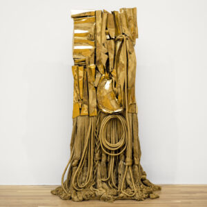 A photo of an abstract, bronze, totemic sculpture. The upper part of the sculpture looks like golden pieces of metal, while the bottom is made of folded, coiled, and braided ropes. It stands on a light wood floor against a white wall. MUST CONTACT HANNAH SHAMBROOM FOR EXTERNAL USAGE PERMISSION