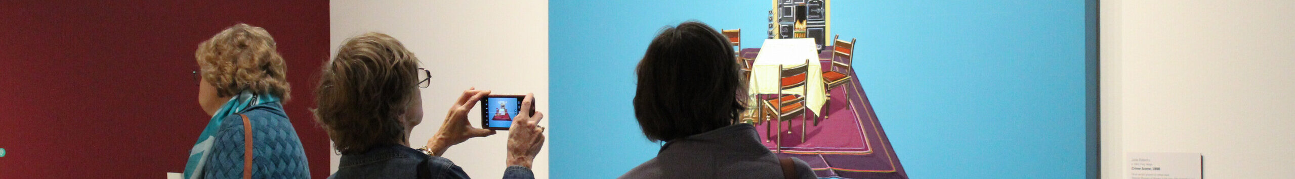 Three museum visitors observe works of art on the wall in a modern gallery. One wall is red and the other is white. One person takes a photo of a work of art with a cell phone.