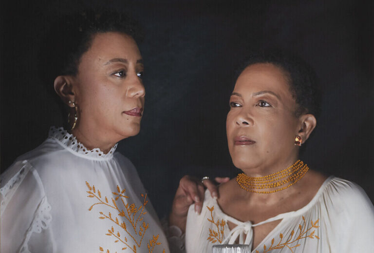 A photograph of two women in embroidered white tops stand behind a table with eggs, orange boxes, pasta in a jar, and a glass pitcher of milk, set against a dark background.