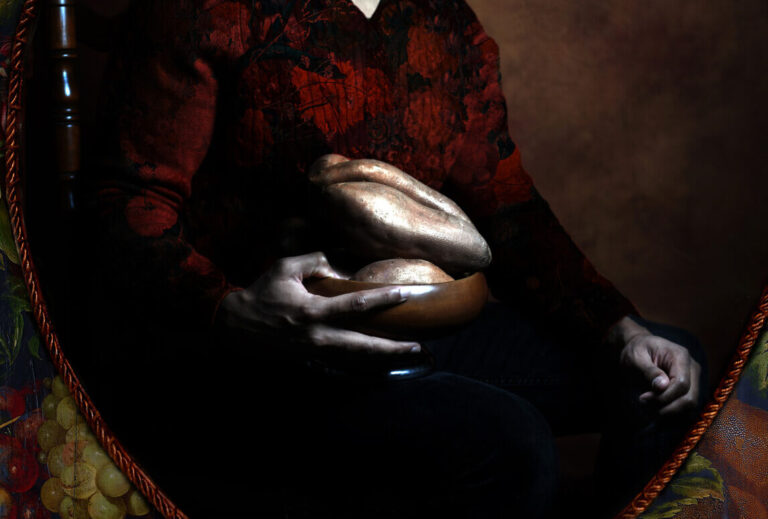 A photograph of a Black man with a beard wearing a red and black shirt, holding a bowl filled with sweet potatoes, and looking towards the right. The background of the photograph is a painted brown color. The photograph is surrounded with braided red trim and a floral background.