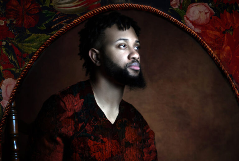 A photograph of a Black man with a beard wearing a red and black shirt, holding a bowl filled with sweet potatoes, and looking towards the right. The background of the photograph is a painted brown color. The photograph is surrounded with braided red trim and a floral background.