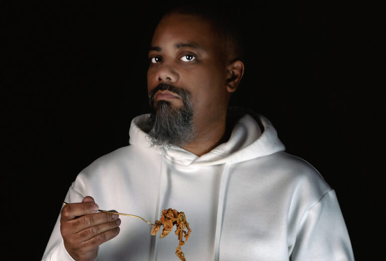 A photograph shows a Black man with facial hair wearing a white hooded sweatshirt. He holds a plate of chitlins in one hand. In the other hand he holds a fork loaded with chitlins.